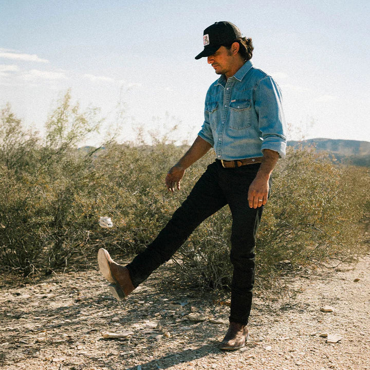 Man in a denim jacket and black cap walking on a dirt path with desert landscape in the background